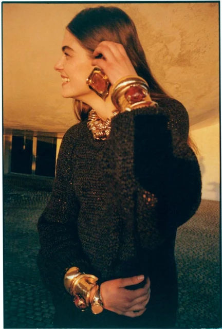 A woman poses in a warm-toned photo, wearing a dark textured top, chunky gold jewelry, and large statement earrings, smiling slightly.