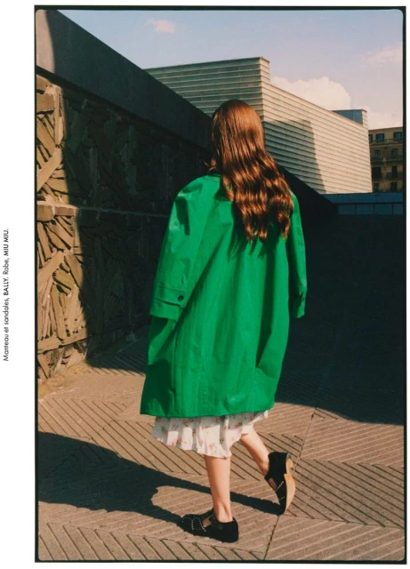 Back view of an adult female model in a green overcoat, black sandals, and a floral dress, walking along a city sidewalk on a sunny day.