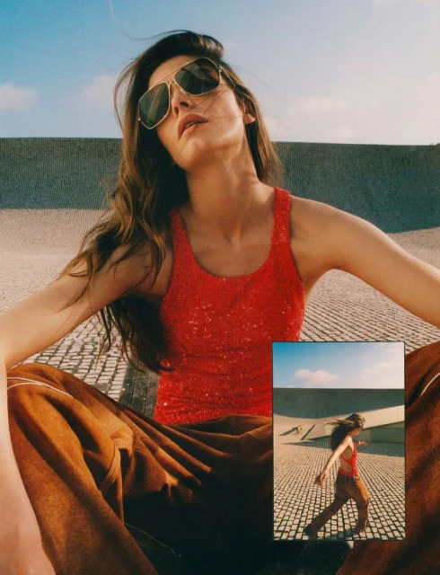 Woman with long wavy hair wears a red top and sunglasses. She sits in an outdoor environment against a textured gray wall.