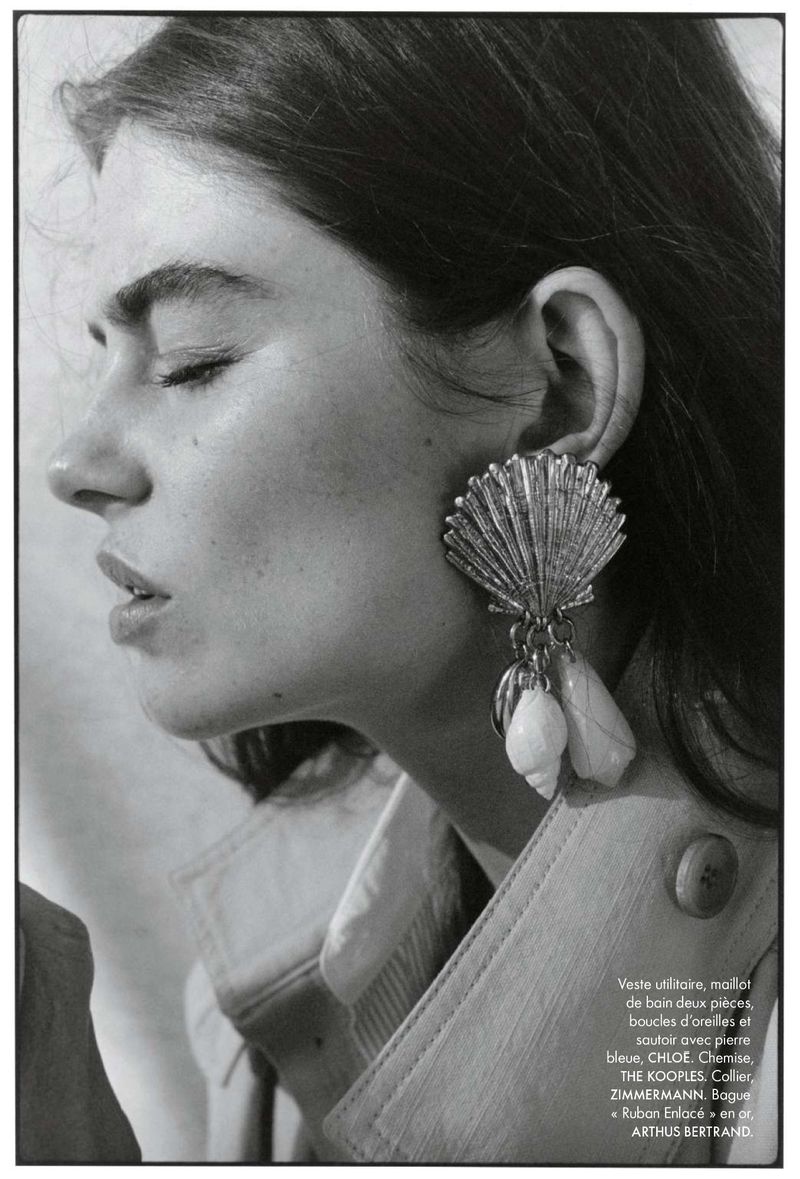 Close-up studio shot: adult woman with eyes closed, wearing a tan jacket and shell-shaped earrings with shells hanging down.