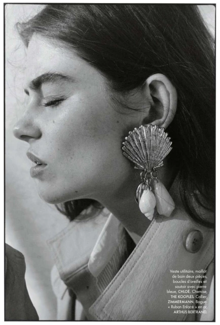 Close-up studio shot: adult woman with eyes closed, wearing a tan jacket and shell-shaped earrings with shells hanging down.