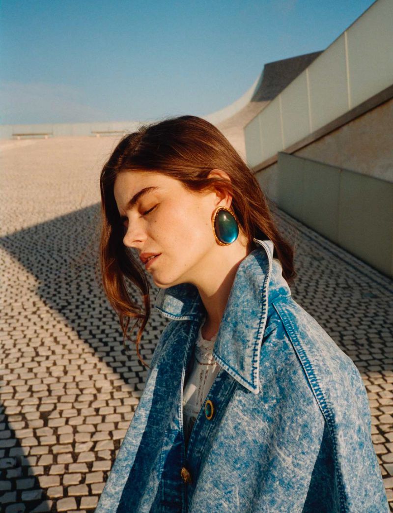 Portrait of a woman with brown hair wearing a denim jacket and large earrings against an abstract architectural background.