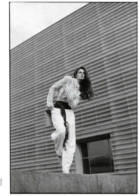 Fashion shot in black and white features a woman in white lace top and pants posing against a modern, horizontally ribbed building.