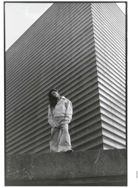 Monochrome shot of a person in relaxed pose, wearing casual clothing, standing against a textured building facade on a cloudy day.