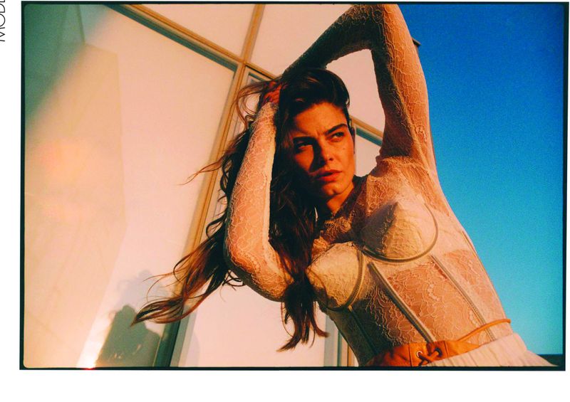 Portrait of a woman in a white lace top with her arms raised, against a blurred blue sky and a building with a glass window.