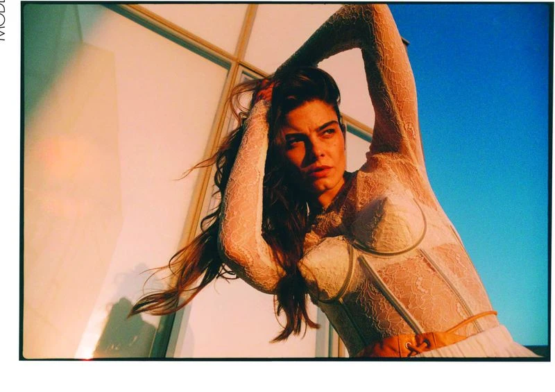 Portrait of a woman in a white lace top with her arms raised, against a blurred blue sky and a building with a glass window.