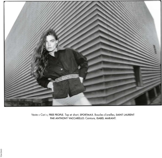 Black and white fashion shot of an adult female wearing a black jacket and ornate belt, posing in front of a corrugated metal building.