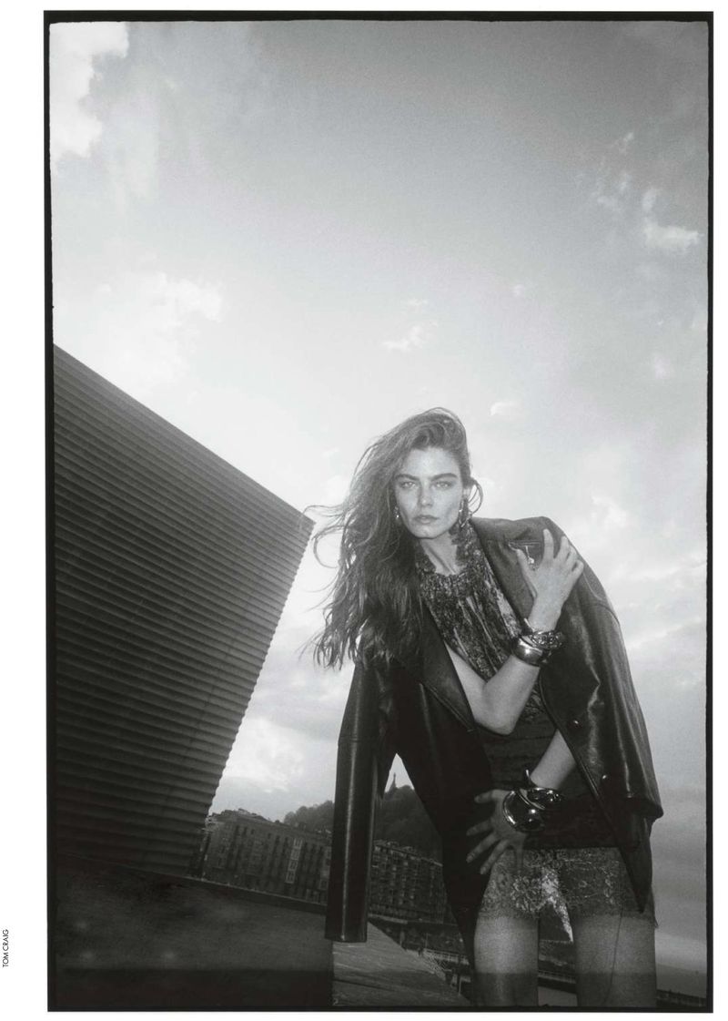 A fashionable woman in a black jacket and patterned dress poses outdoors against a bright sky and modern architectural structure in a monochrome image.