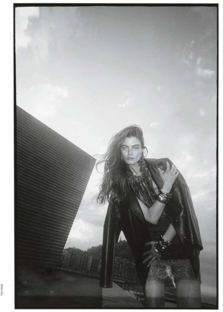 A fashionable woman in a black jacket and patterned dress poses outdoors against a bright sky and modern architectural structure in a monochrome image.