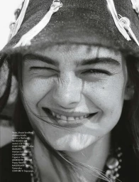 Close-up black and white portrait of a smiling female model wearing a textured hat with ribbons and a necklace for Elle France.