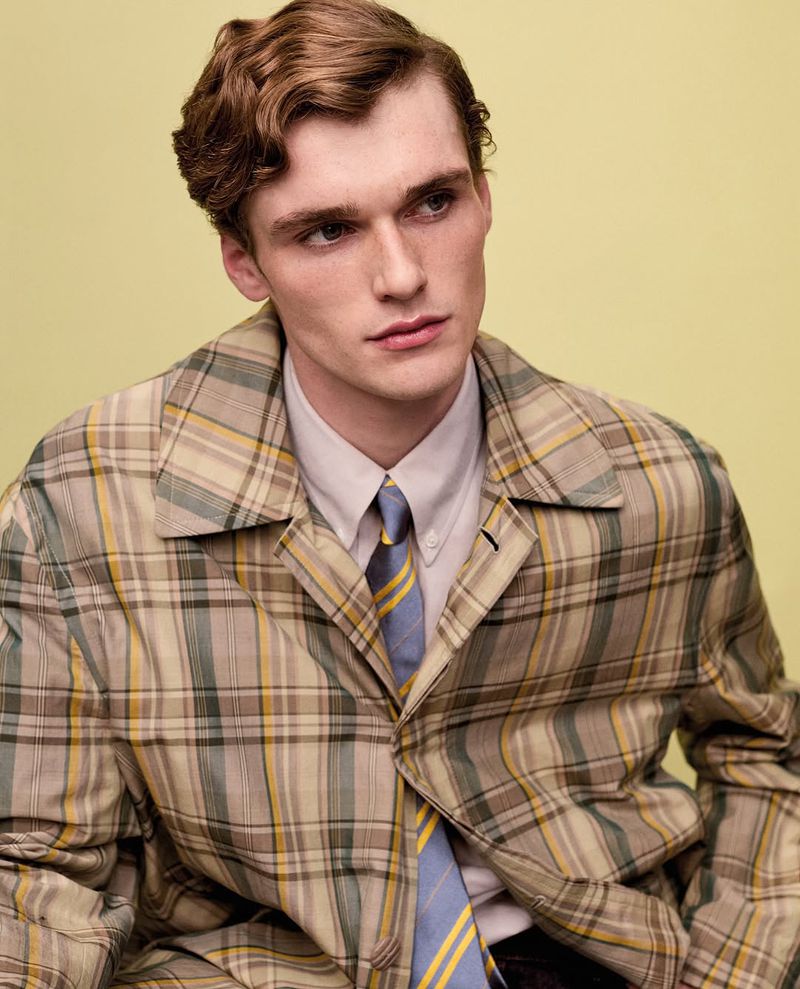 Close-up portrait of an adult male with wavy auburn hair wearing a tan, blue, and yellow plaid jacket, white shirt, and light blue tie.