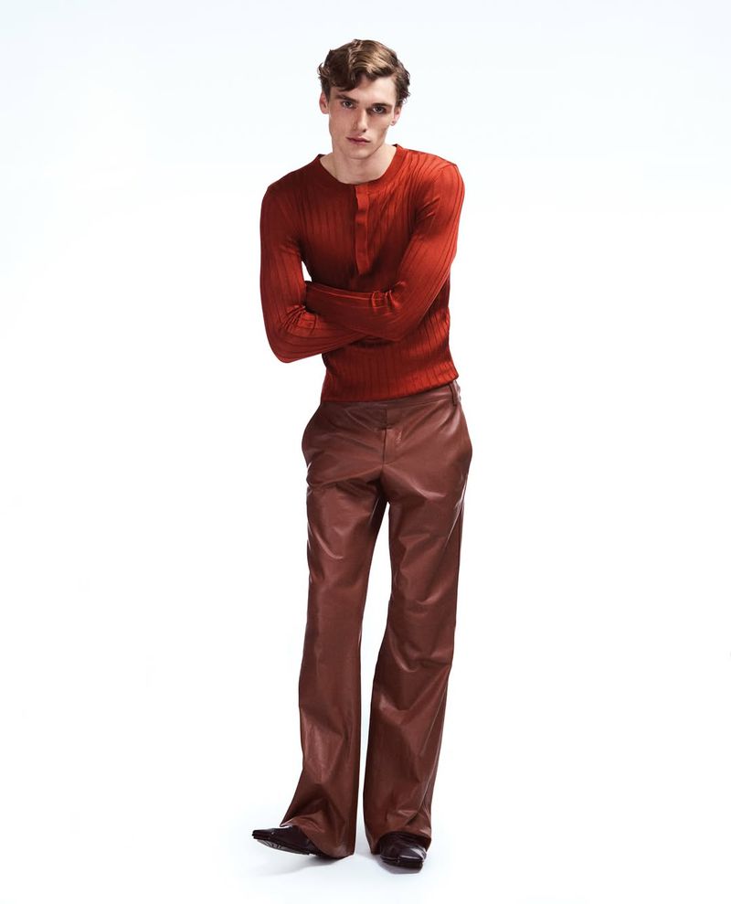 Full-length portrait of an adult male fashion model with folded arms wearing a rust-colored sweater and shiny brown leather trousers in a studio.