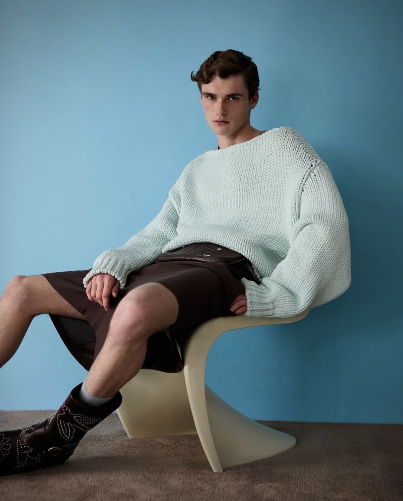 Man poses in studio wearing a light blue sweater, brown shorts, and cowboy boots, seated on a chair against a blue wall.