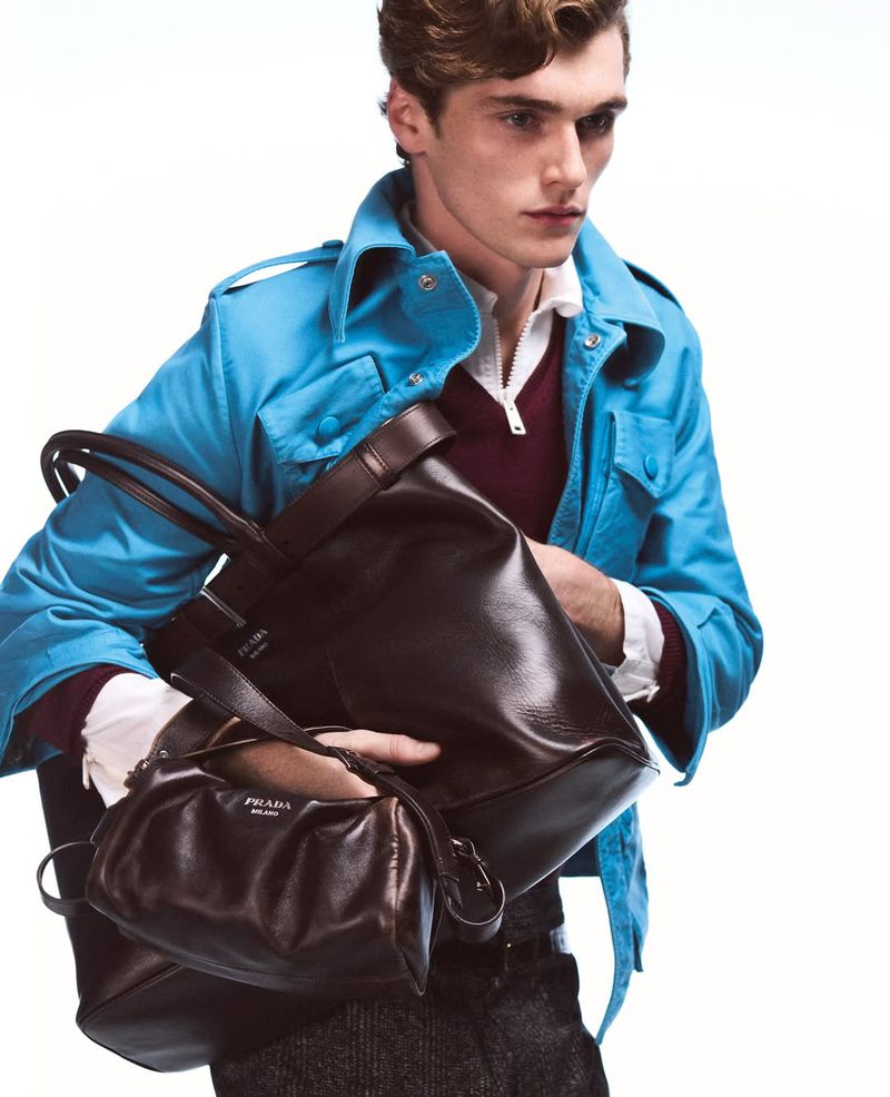 Male model poses in a blue jacket and dark trousers, holding a black leather handbag and small purse against a white background.