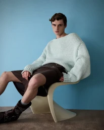 Model seated on a curved white chair wearing a mint green knit sweater and brown shorts against a blue background.