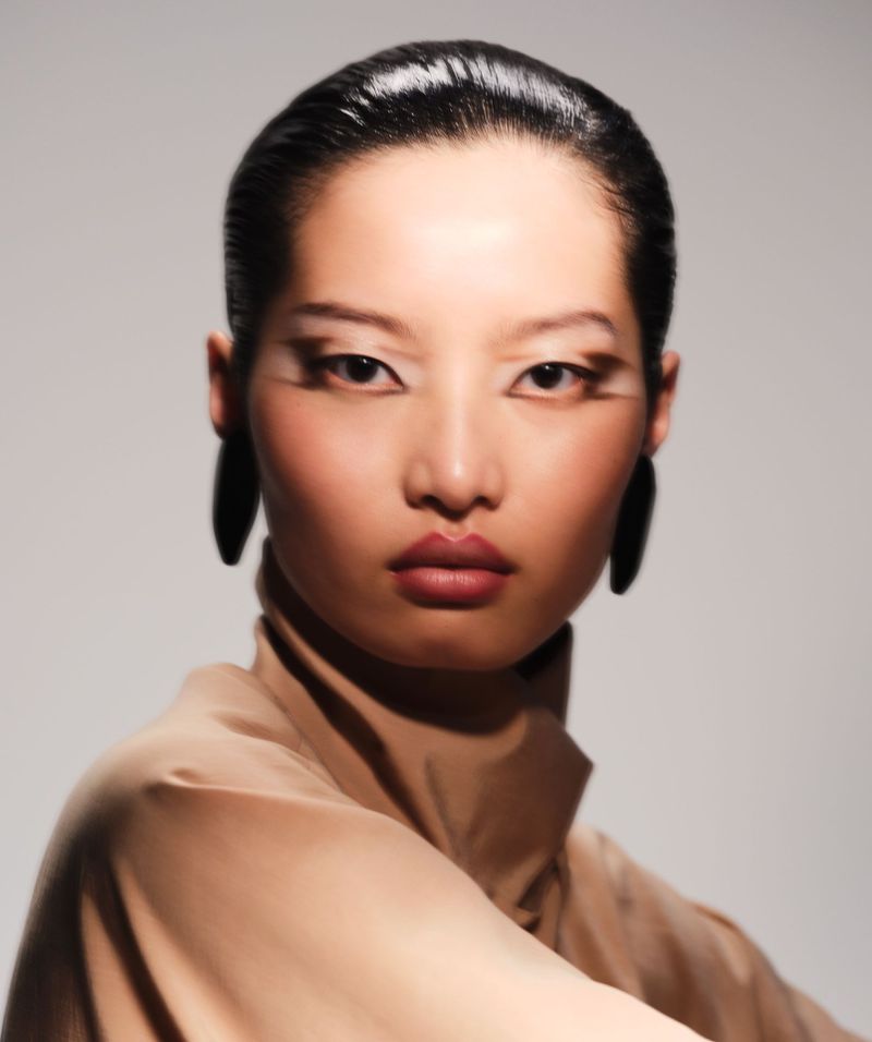 Fashion portrait of a woman with dark hair, tan turtleneck, statement earrings, and dramatic eye makeup in a photo studio.