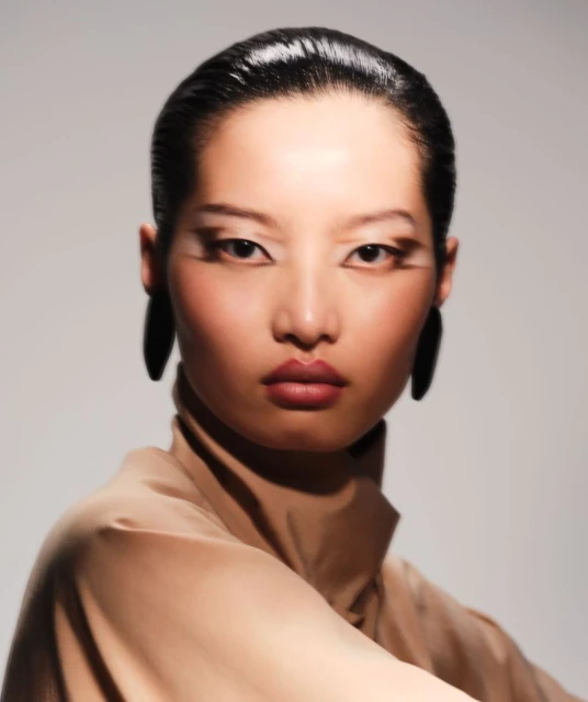 Fashion portrait of a woman with dark hair, tan turtleneck, statement earrings, and dramatic eye makeup in a photo studio.