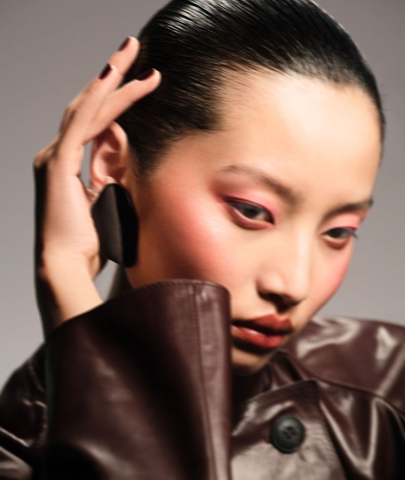 Close-up of an Asian woman with sleek hair and brown leather jacket, striking a pose with hand near her head, set against a grey background.