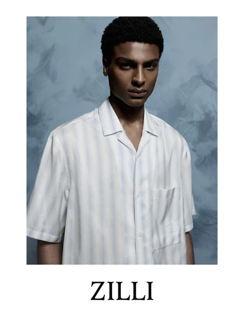 Dark-skinned adult male poses indoors wearing a striped collared short-sleeve shirt in a fashion-style portrait against a blue background.
