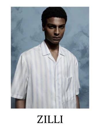 Dark-skinned adult male poses indoors wearing a striped collared short-sleeve shirt in a fashion-style portrait against a blue background.