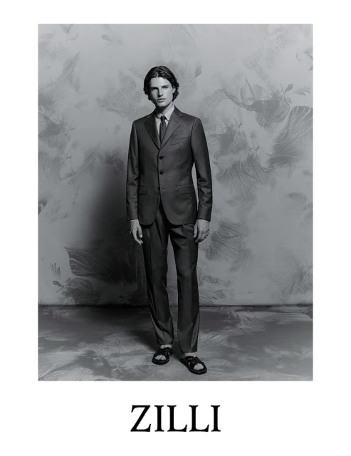 Monochrome full shot of a male fashion model wearing a suit with dress shoes against a textured backdrop, advertising Zilli.