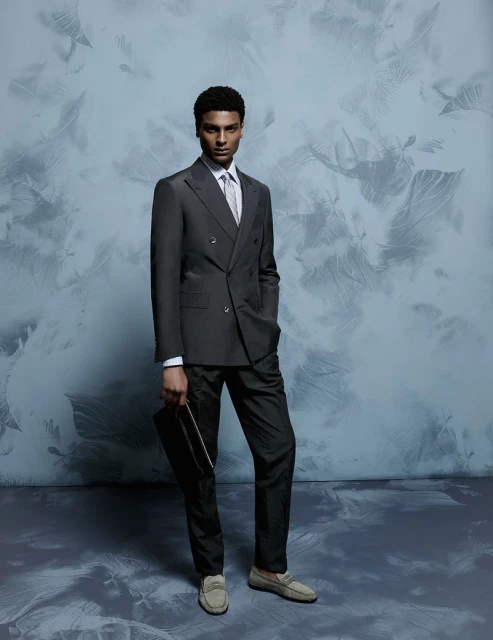 Full-length portrait of a young Black man in a double-breasted grey suit, white shirt, and loafers, holding a clutch against a light blue background.