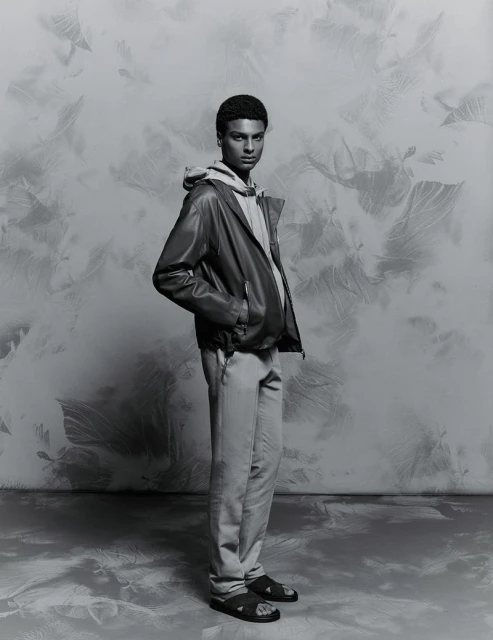 Monochrome studio portrait of a young adult with an afro hairstyle, posing in a leather jacket, hoodie, and pants against a mottled backdrop.