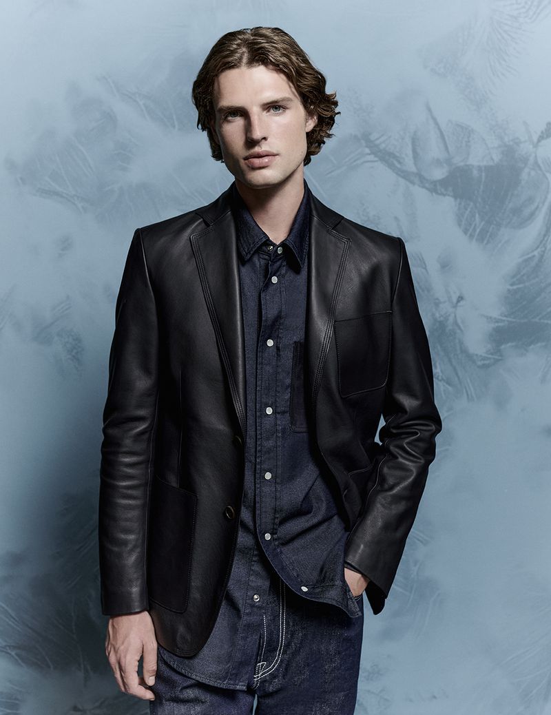 Man in a dark blue shirt and a black leather jacket poses against a light blue background in a fashion-style portrait, indoors.