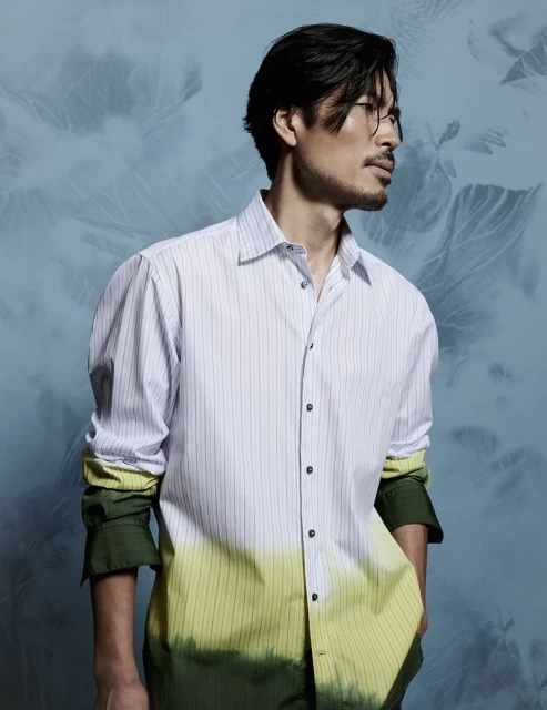 Studio shot of an adult male model wearing a striped button-down shirt with an ombre green/yellow dye effect, posing against a blue background.
