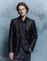 A male model wearing a black leather blazer over a dark blue button-down shirt and denim jeans against a textured blue backdrop.