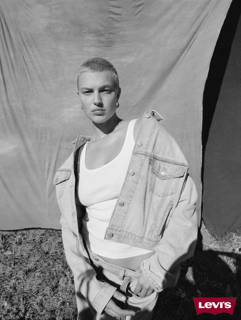 Black and white fashion portrait of a woman with a buzzcut wearing a denim jacket, a white tank top and jeans in front of a draped backdrop.