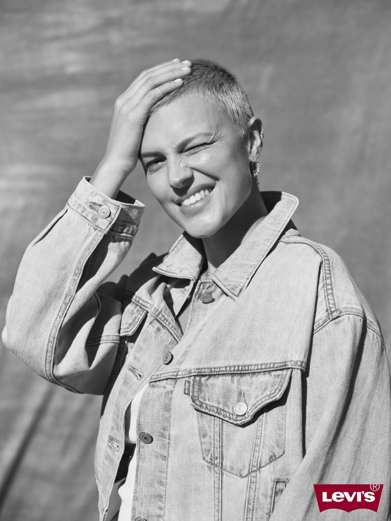 Close-up black and white portrait of a smiling woman with short hair wearing a denim jacket, against a grey backdrop with the Levi's logo.