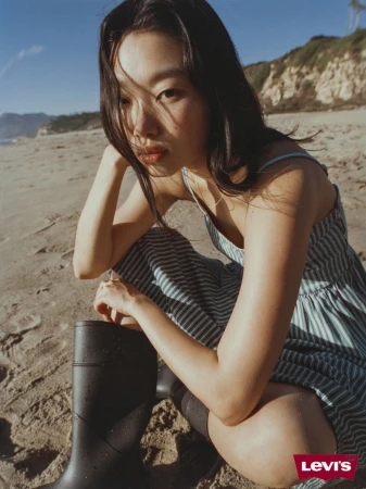 An Asian woman poses on a sandy beach in a striped sundress and rain boots, bathed in soft sunlight with a coastal backdrop.