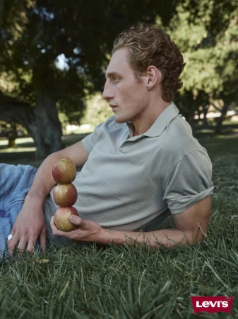 Man lying on grass in a park holding stacked apples. He wears a grey collared shirt with trees and sky visible in the background.