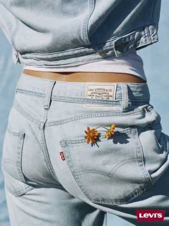 Close-up studio shot of a person wearing a denim jacket and jeans with flowers in the pocket against a light blue backdrop.