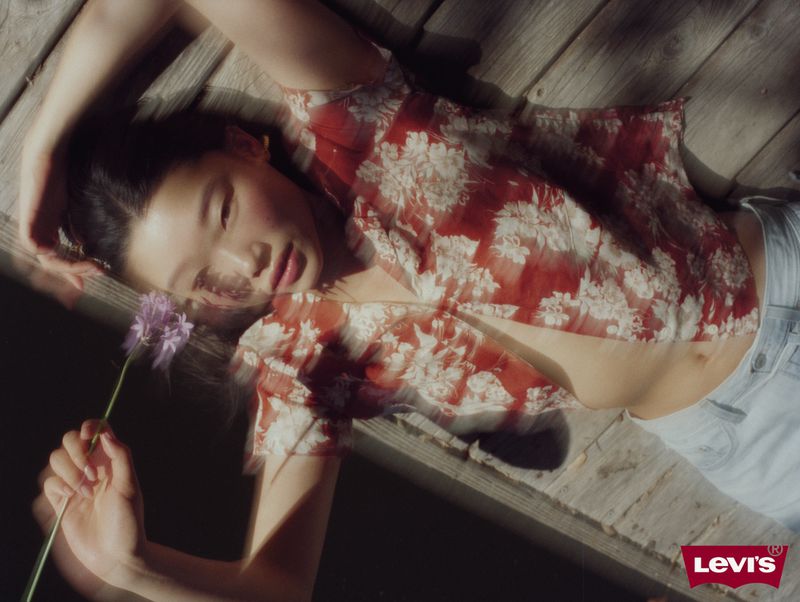 Young woman lies on a wooden deck, holding a purple flower in her hand, wearing a floral blouse and light-wash jeans, lit by natural light.