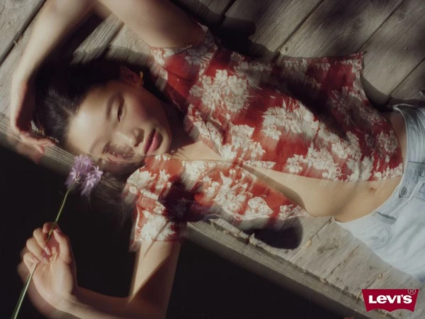 Young woman lies on a wooden deck, holding a purple flower in her hand, wearing a floral blouse and light-wash jeans, lit by natural light.