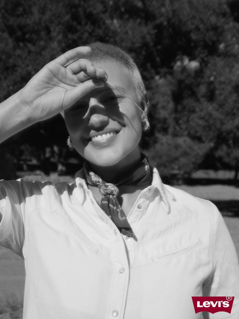 Close-up black and white portrait of a smiling woman with short hair, wearing a collared shirt and bandana with greenery in the background.