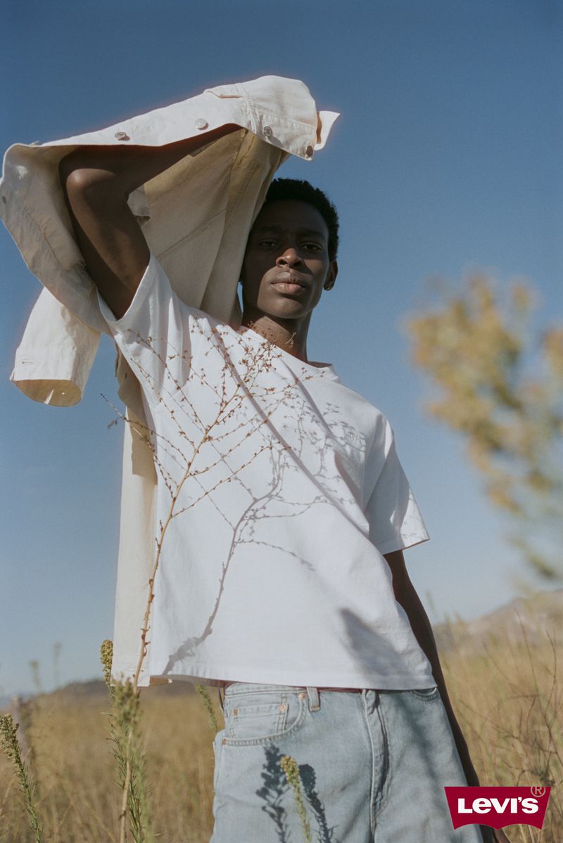 Outdoor fashion portrait: A young Black male model in jeans and a white tee poses with a jacket, set against a sunlit field and sky.