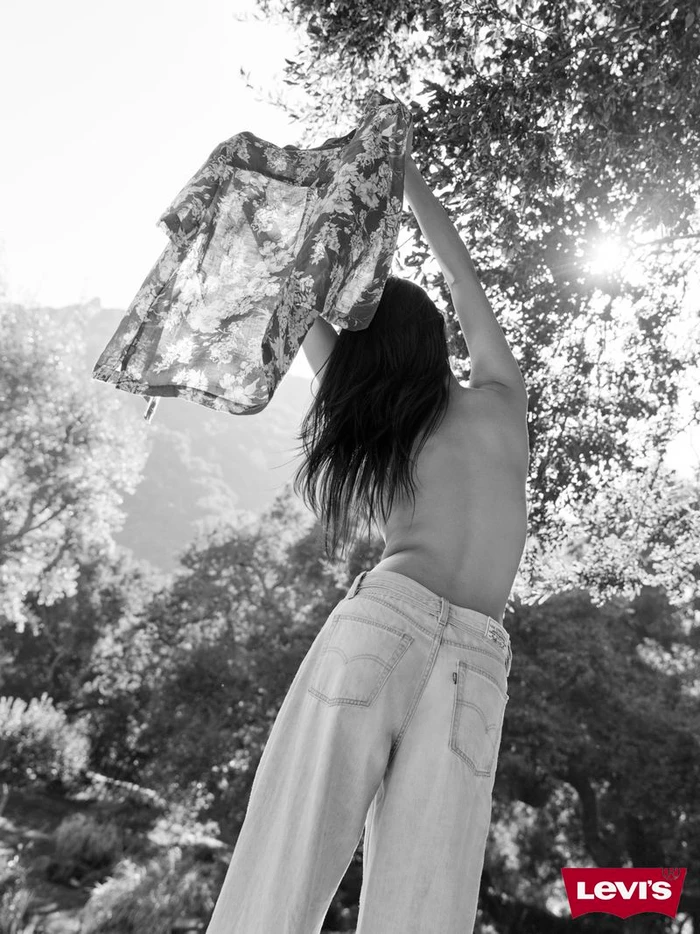 Black and white portrait of a woman lifting a floral shirt outdoors, wearing denim jeans in sunlight