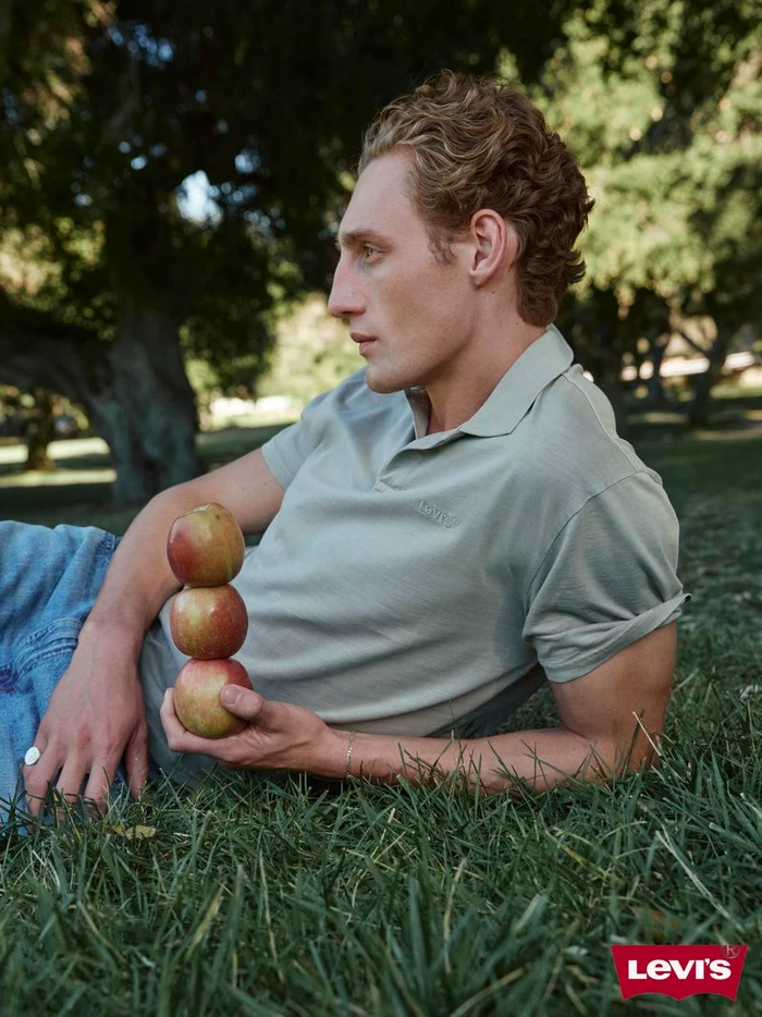 Man with curly blonde hair lying on grass holding a vertical stack of three red apples outdoors