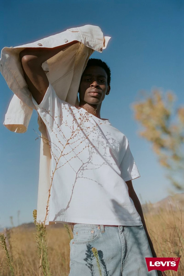 Black man holding a white denim jacket over his shoulder in a field under clear blue sky for Levi's Spring 2025 campaign.