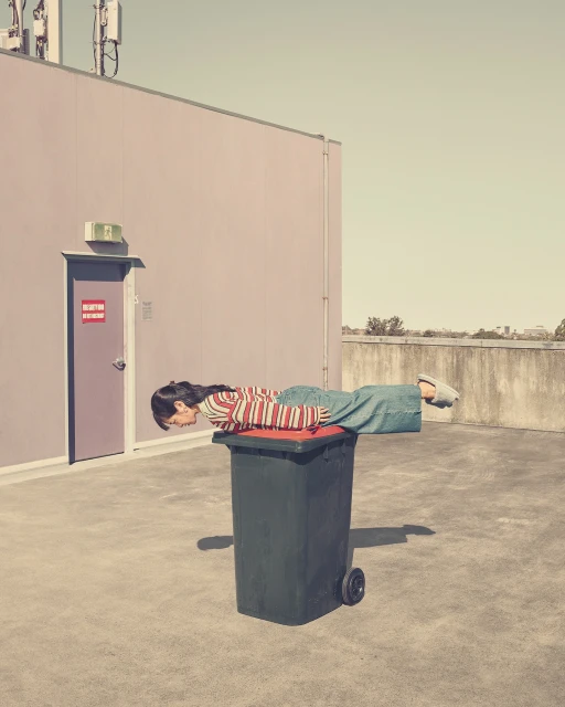 Woman planking on a trash can outdoors with building and sky visible. Unusual pose, exercise on a rooftop in casual clothing, daytime.