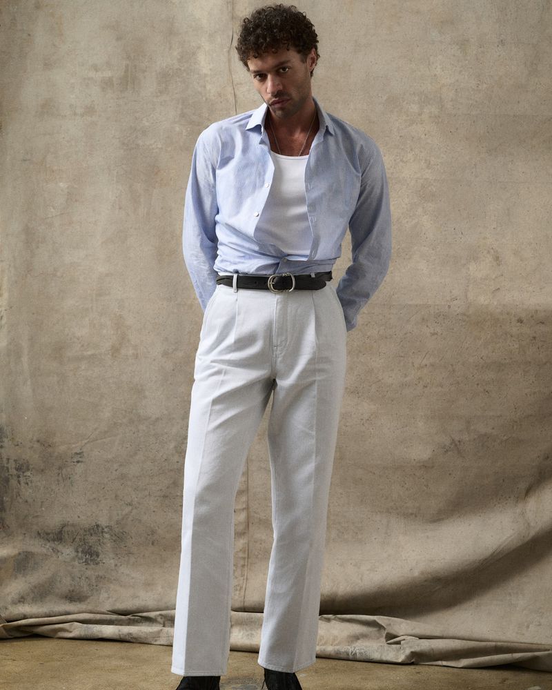 Man with curly hair in blue shirt, white undershirt, and white pants poses against a neutral backdrop in a studio setting.