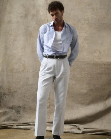 Man with curly hair in blue shirt, white undershirt, and white pants poses against a neutral backdrop in a studio setting.