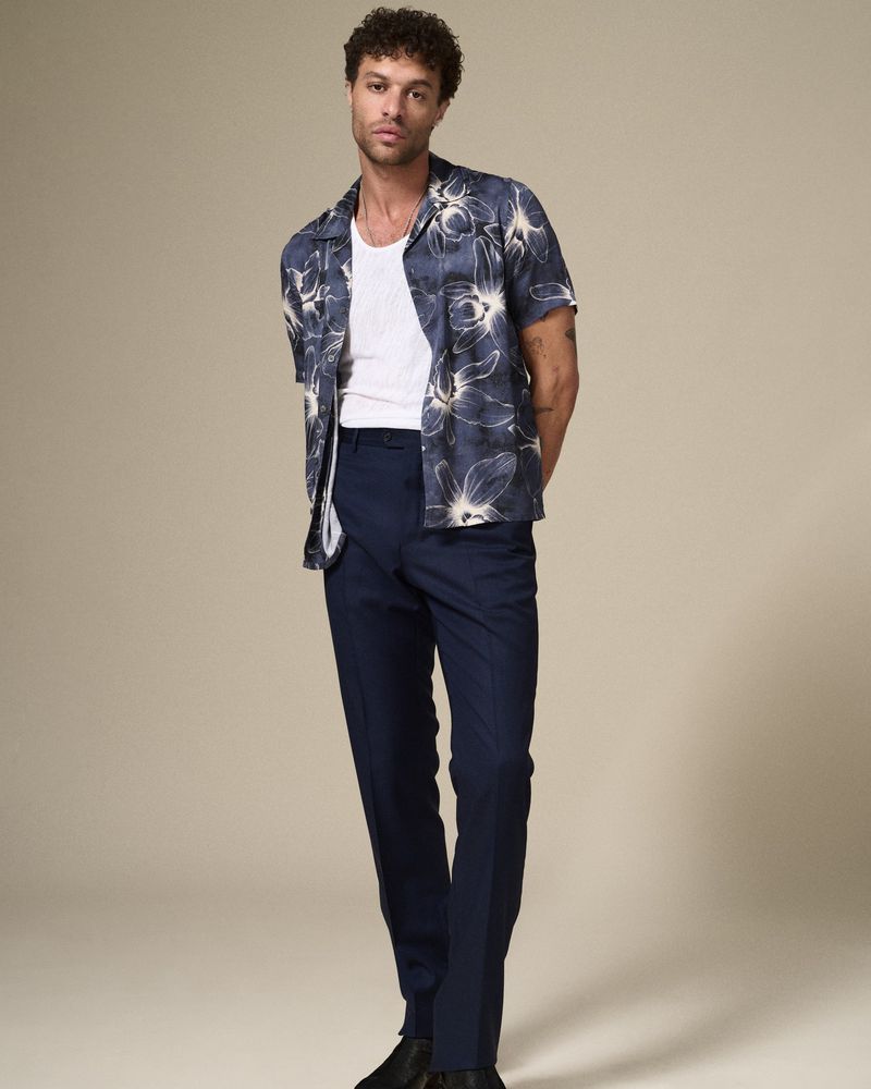 Full-length portrait of a young adult male in floral short-sleeved shirt, white undershirt, and blue slacks, posing in a neutral studio.