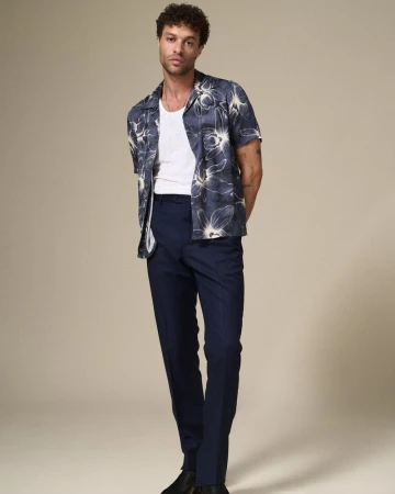 Full-length portrait of a young adult male in floral short-sleeved shirt, white undershirt, and blue slacks, posing in a neutral studio.