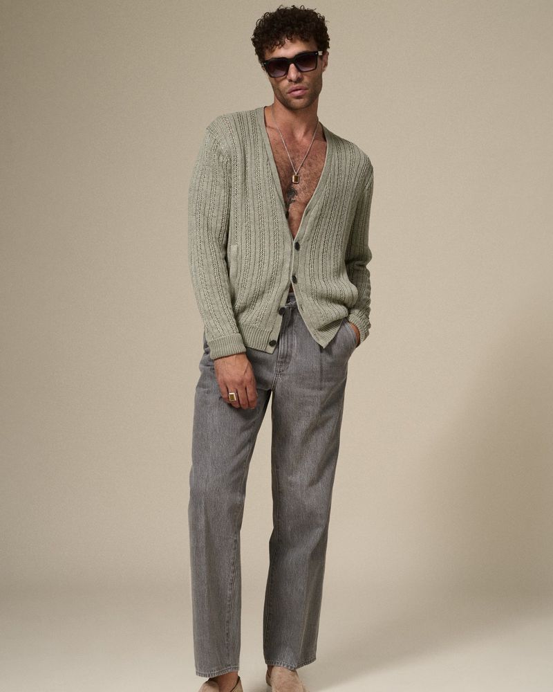Portrait of adult male model wearing sunglasses, a sage cardigan, and grey striped pants standing against a neutral studio backdrop.