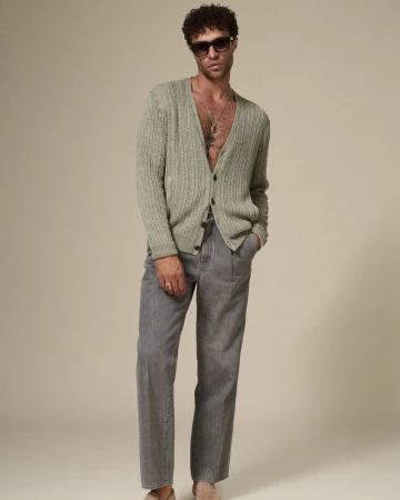 Portrait of adult male model wearing sunglasses, a sage cardigan, and grey striped pants standing against a neutral studio backdrop.