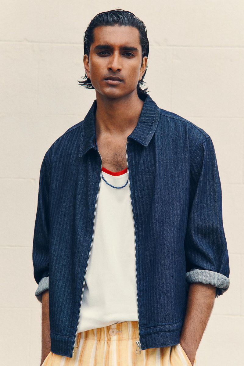 Portrait of a dark-haired man wearing a blue pinstripe zip jacket over a white t-shirt with yellow striped pants against a neutral wall.